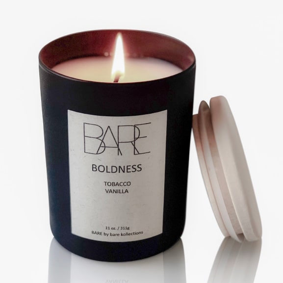 Boldness scented candle with open flame in a black jar. A wooden lid is leaned on it. White label says Bare Boldness Tobacco Vanilla.