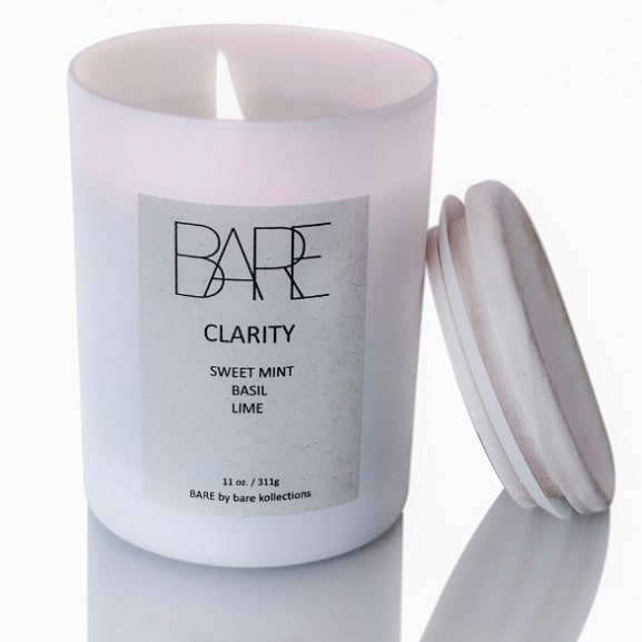 Clarity scented candle with open flame in a white jar. A wooden lid is leaned on it. White label says BARE, Sweet Mint, Basil, Lime.