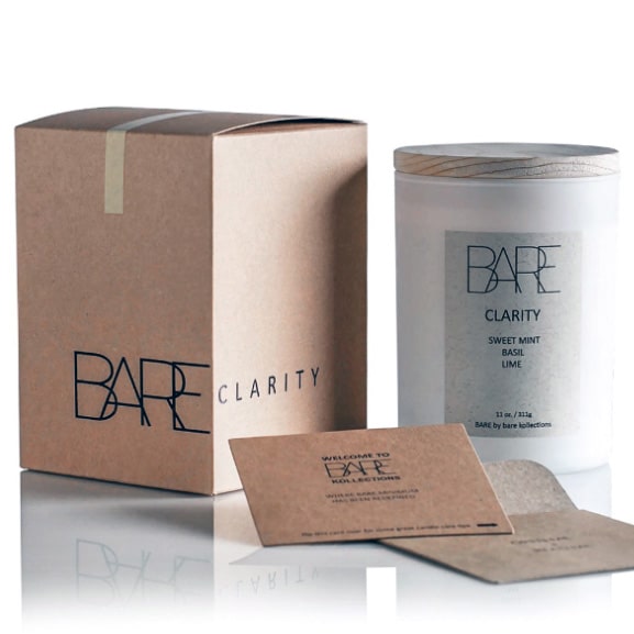 White background. Bare recycable brown box with Bare logo and Clarity typography, turned sideways. Next to it, on the right side is a scented candle in a closed white jar with beige Clarity label on it. In front are care instructions on a cardboard recycable material.