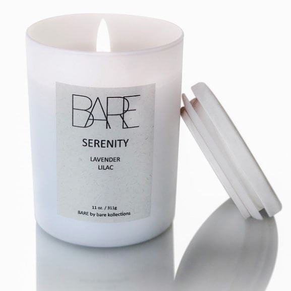 Serenity scented candle with open flame in a white jar. A wooden lid is leaned on it. White label says BARE, Lavender, Lilac.