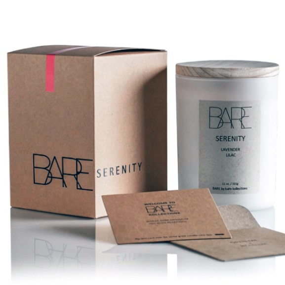 White background. Bare recycable brown box with Bare logo and Serenity typography, turned sideways. Next to it, on the right side is a scented candle in a closed white jar with beige Serenity label on it. In front are care instructions on a cardboard recycable material. 